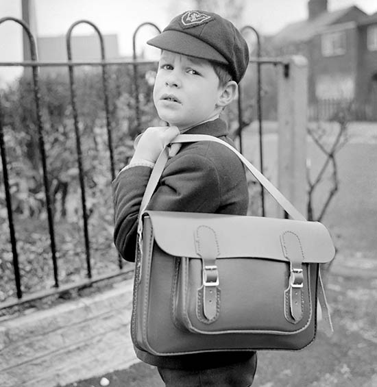 A child stood by railings in school uniform and holding a satchel