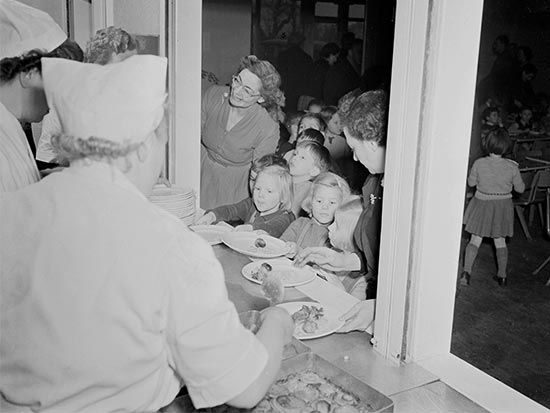 Dinner ladies serving lunch to a group of children at a hatch